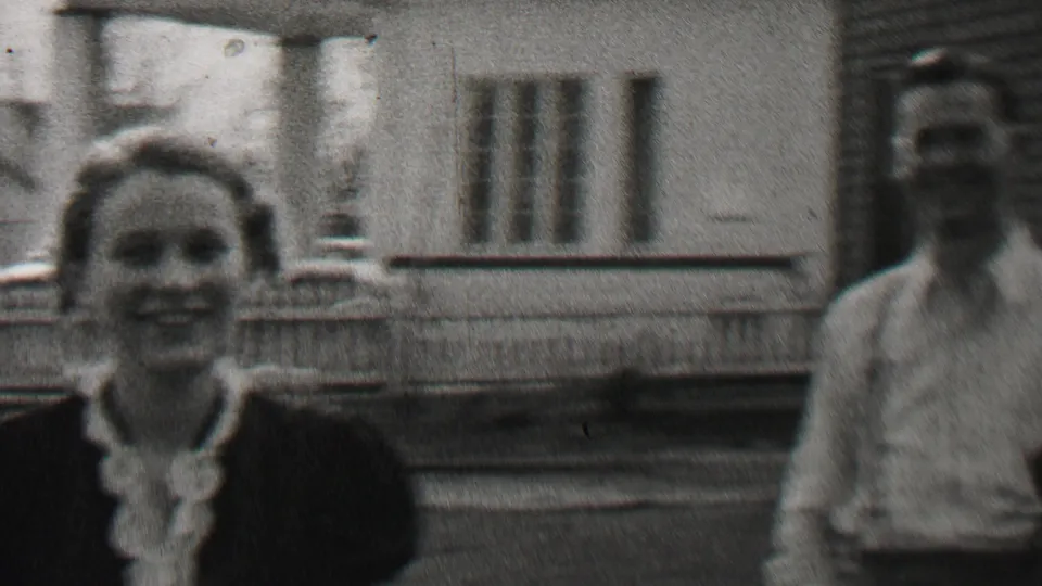 A woman in a dark dress with a light collar smiles toward the camera as the man in a light shirt stands partly cropped at the edge of the Beachwood porch scene.