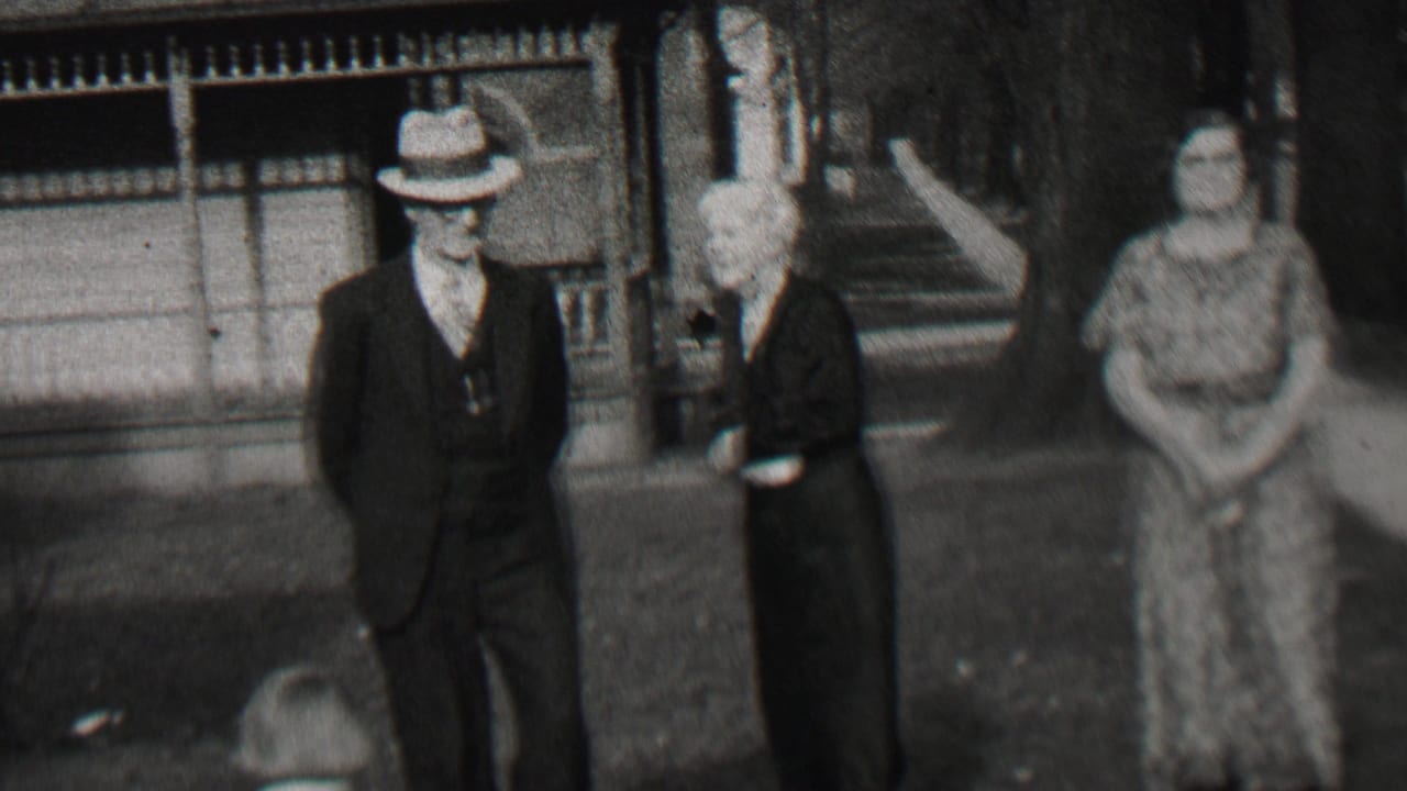 In Beachwood, New Jersey in 1937, a suited man in a fedora stands near two women in dark and patterned dresses on a tree-lined residential sidewalk.