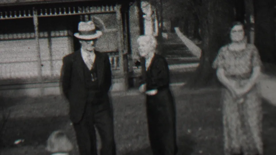 In Beachwood, New Jersey in 1937, a suited man in a fedora stands near two women in dark and patterned dresses on a tree-lined residential sidewalk.