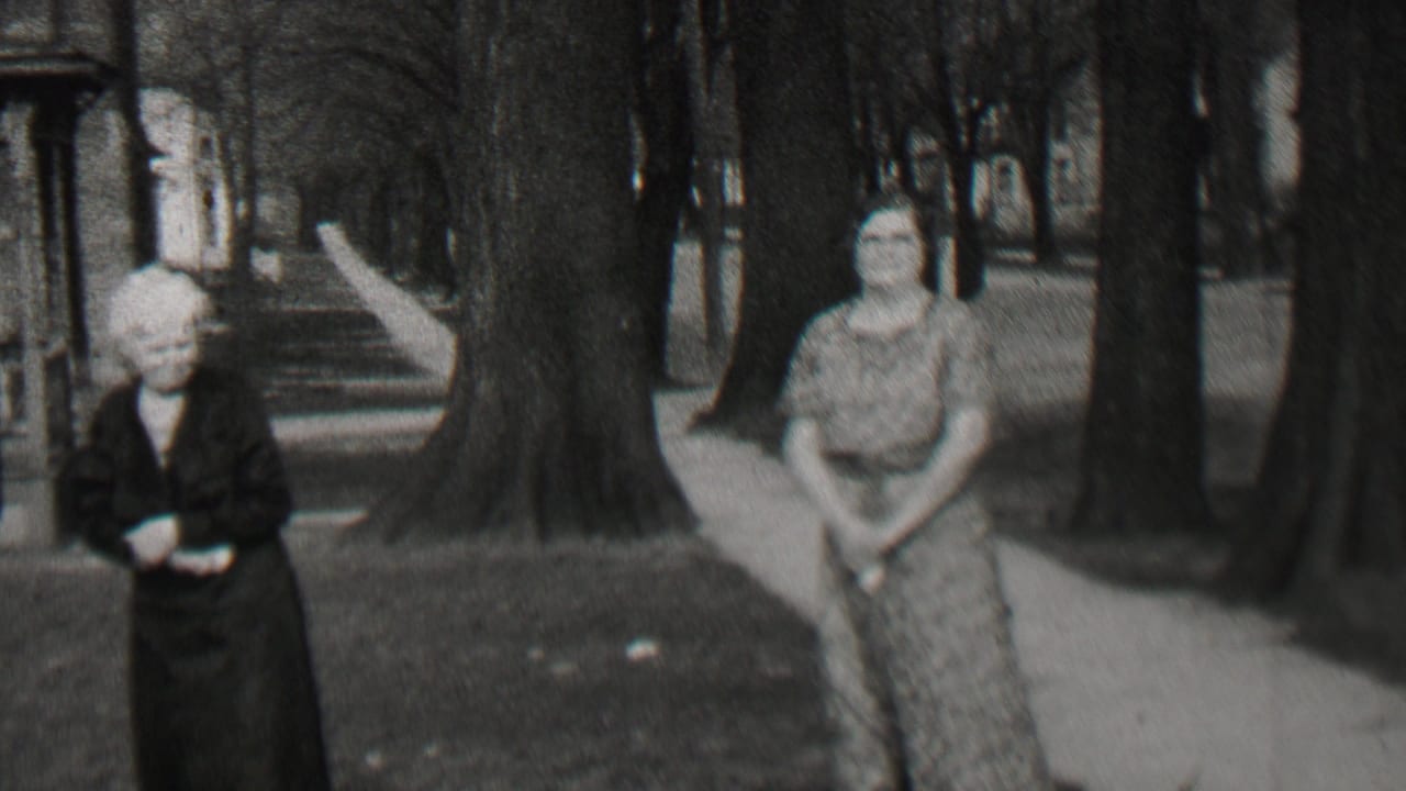 Two women stand apart on grass and sidewalk near large trees and porch steps.