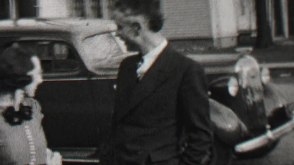 A suited man stands next to a dark 1930s automobile in the Beachwood, New Jersey home-movie footage, with adults and a child-sized figure nearby.
