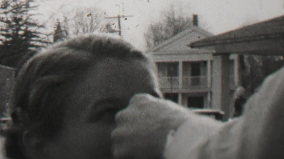 Two women appear close to the camera in a 1937 Beachwood, New Jersey residential yard or street setting, with houses, porch railings, bare trees, and a picket fence behind them.