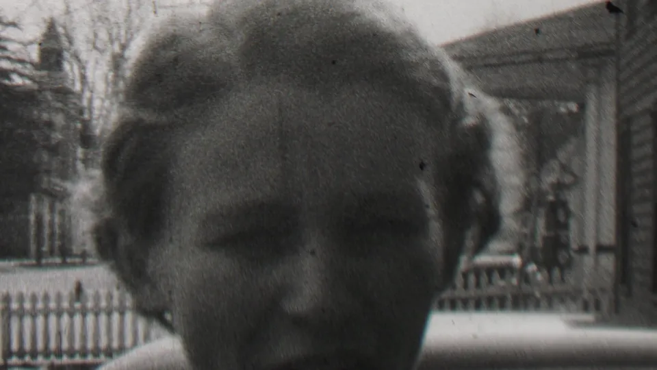 A woman is framed close to the camera in a 1937 Beachwood, New Jersey home-movie scene, with a clapboard house, porch, railings, and fence visible in the background.