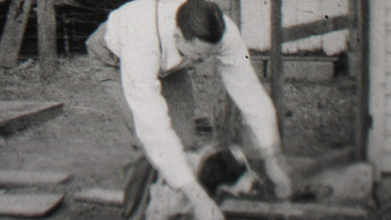 In 1937 Beachwood, New Jersey home-movie footage, an adult man in a white shirt and suspenders bends down toward a small black-and-white dog beside a wooden residential structure.