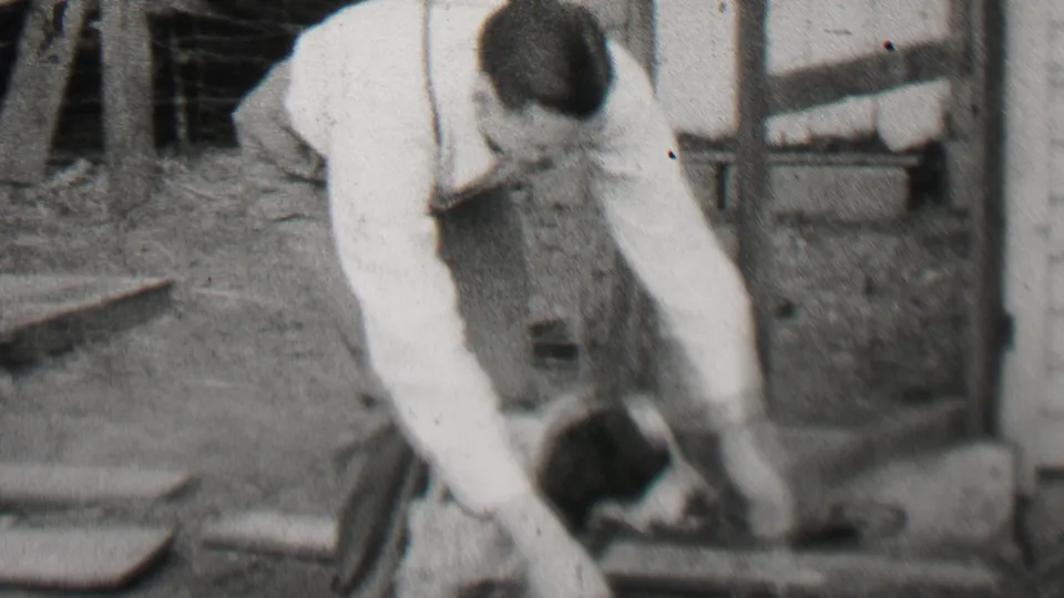 In 1937 Beachwood, New Jersey home-movie footage, an adult man in a white shirt and suspenders bends down toward a small black-and-white dog beside a wooden residential structure.