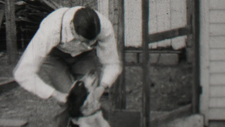 A crouching adult man in suspenders reaches toward the small dog in a grainy 1937 Beachwood backyard home-movie frame, with wood siding and planks nearby.