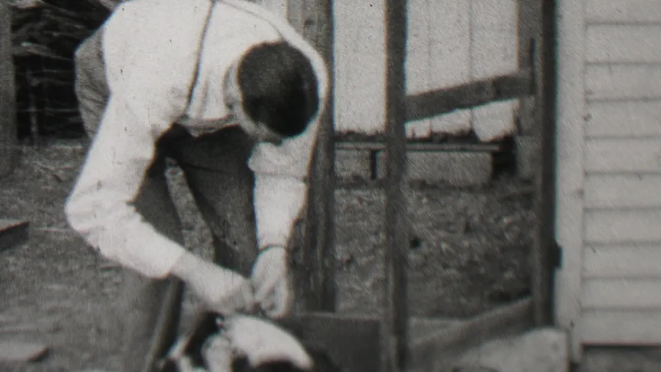 The man bends over the small dog near house siding and a porch support, continuing the close backyard interaction in the 1937 Beachwood home movie.