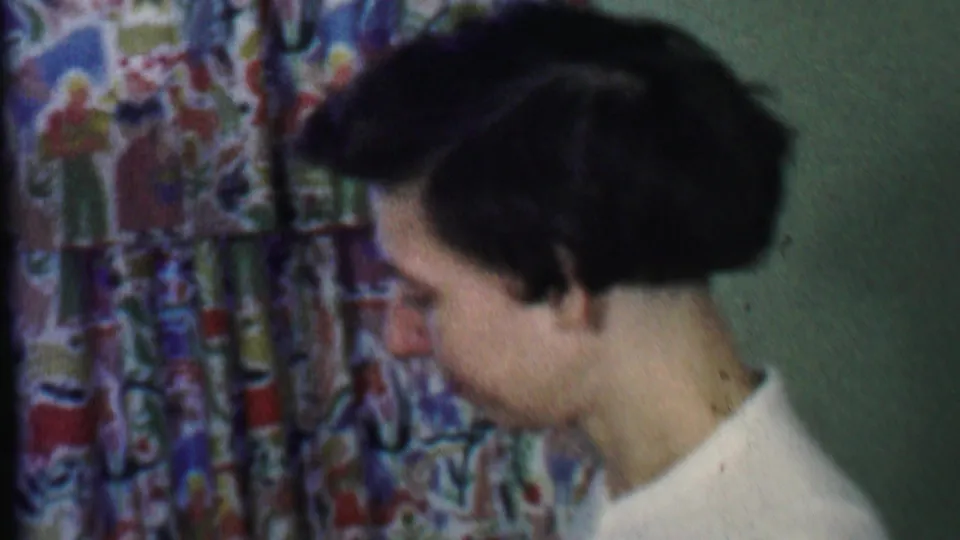 In this 1959 Des Moines home-movie frame, a woman in a white shirt is seen in profile near colorful patterned curtains inside a private interior.