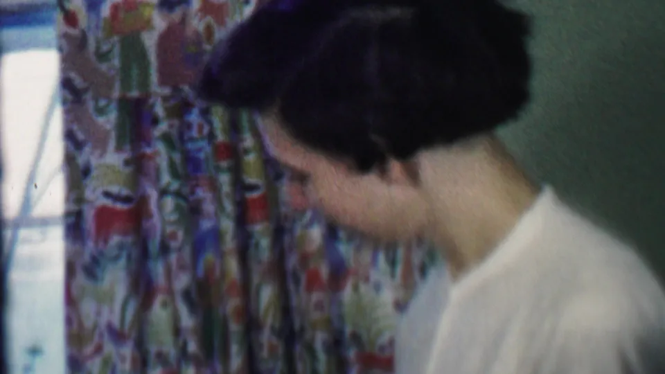 A close 8mm frame shows the woman looking down as if concentrating on a task, with patterned curtains and a bright window edge behind her.