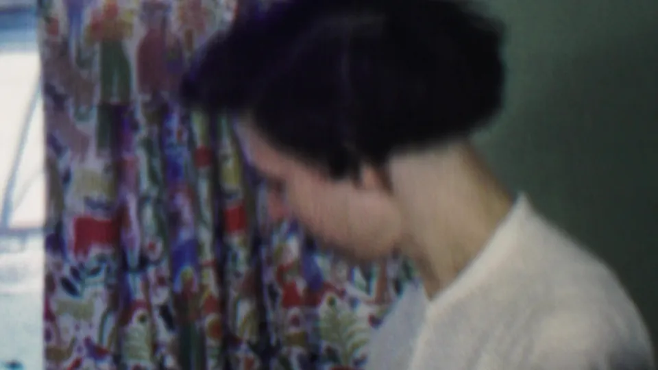 The final frame keeps the woman in close side view as she looks downward near colorful curtains, preserving a quiet domestic moment from the 1959 Des Moines home movie.