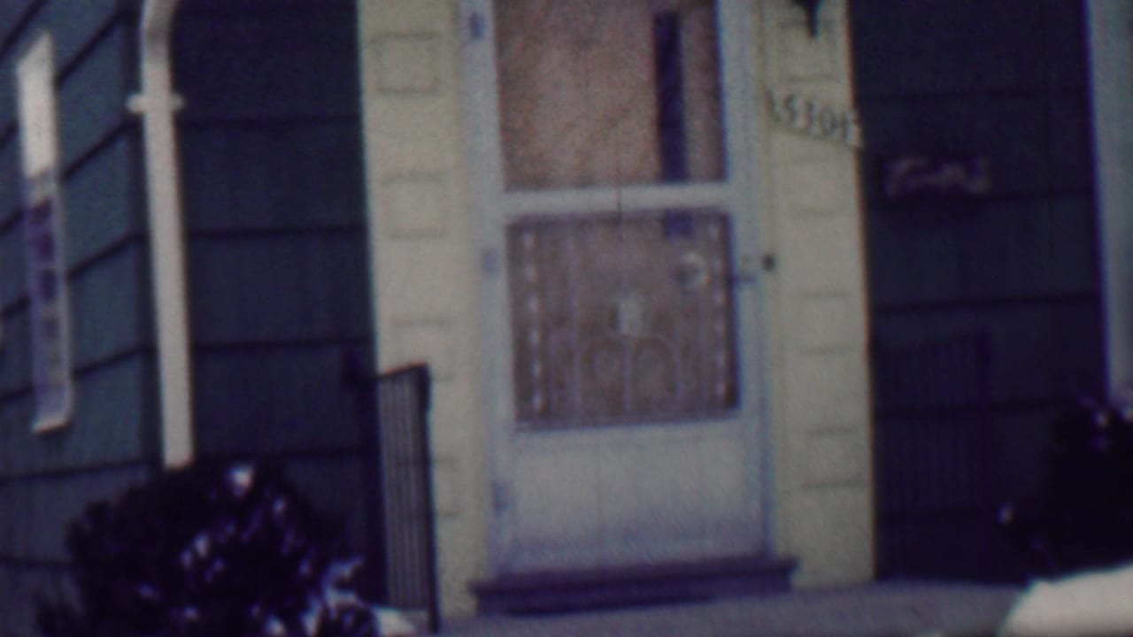 1958 Grand Rapids home-movie frame showing a snowy residential front entrance with dark siding, a light decorative door, small railings, and shrubs in the foreground.