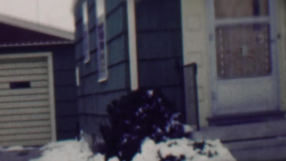 1958 Grand Rapids home-movie frame showing part of a residential facade with a garage door, dark siding, a light entrance area, snow, and a foreground shrub.