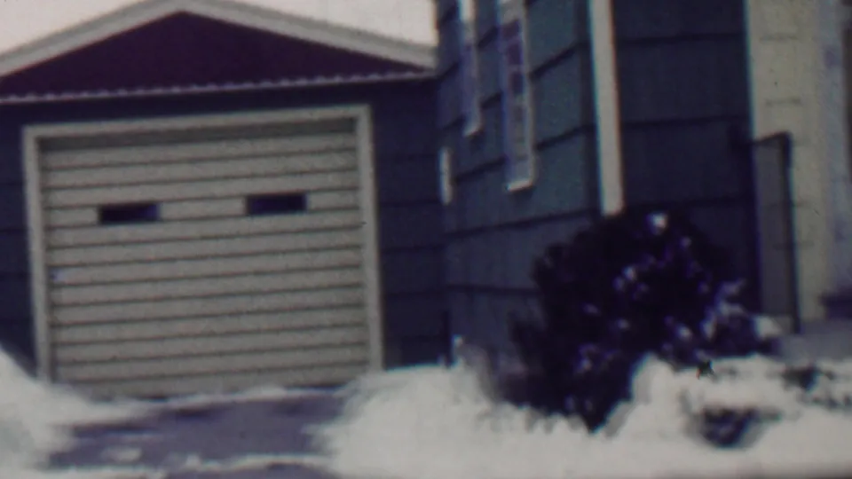 1958 Grand Rapids frame showing a garage door and adjacent residential entrance area, with dark siding, a small railing, snow-covered shrubs, and winter ground cover.