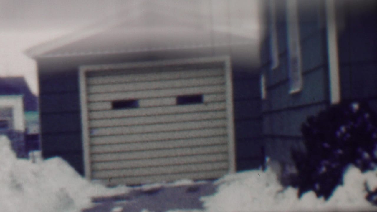 1958 Grand Rapids home-movie frame showing a snowy driveway, detached garage, dark house siding, and high snowbanks in a residential neighborhood.