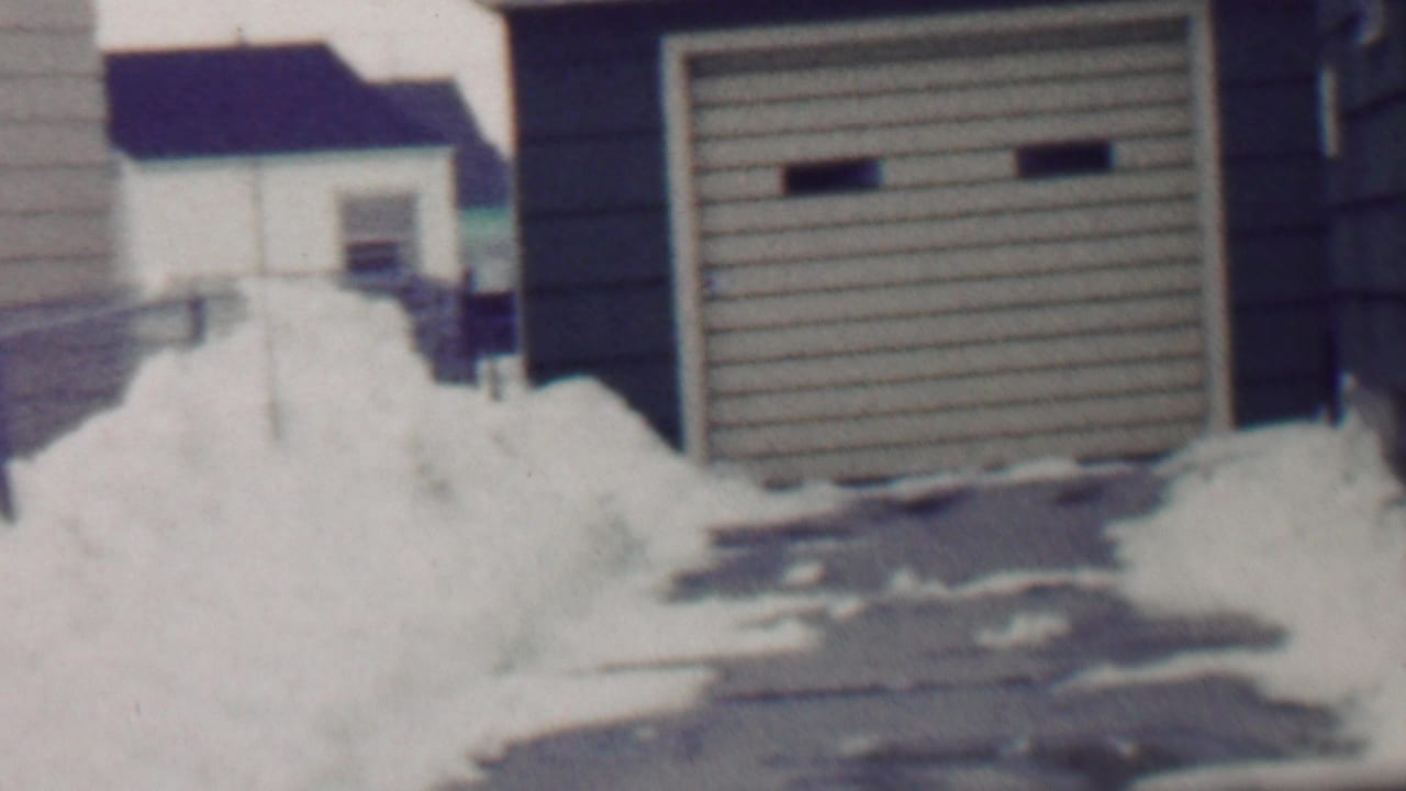1958 Grand Rapids frame with a detached garage, snow piled along the driveway, and neighboring houses partly visible beyond the yard.