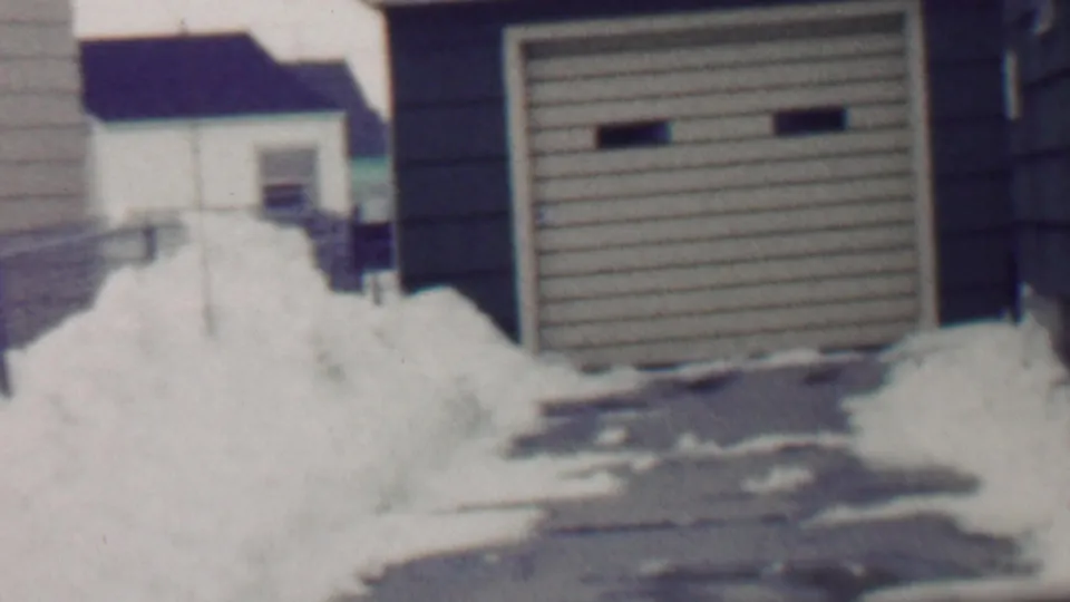 1958 Grand Rapids frame with a detached garage, snow piled along the driveway, and neighboring houses partly visible beyond the yard.