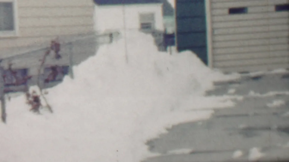 Grand Rapids winter home-movie frame from 1958 showing the side of a house, a driveway edge, bright snowbanks, and a partially visible neighboring structure.