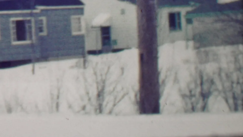 1958 Grand Rapids home-movie frame showing a snowy residential view with houses, a utility pole, bare shrubs, snowbanks, and a driveway in winter.