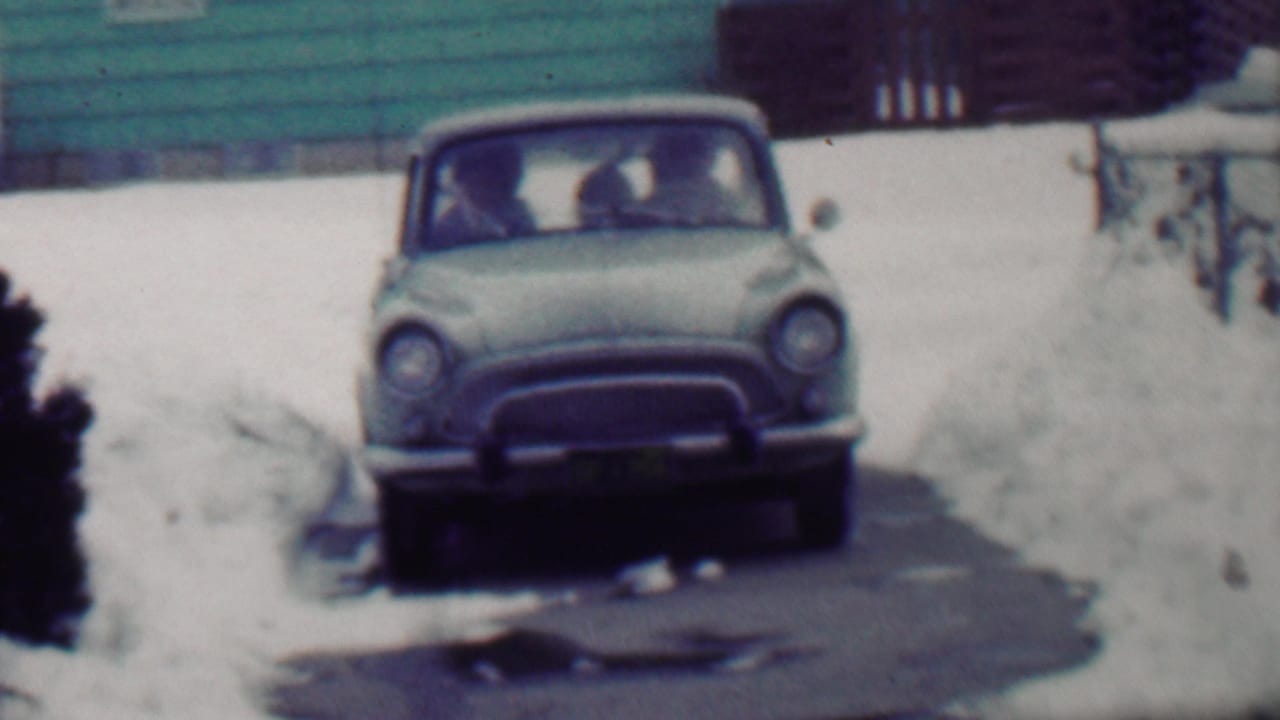 A light-colored mid-century automobile faces the camera on a snow-covered residential driveway in Grand Rapids, Michigan, with passengers faintly visible through the windshield.