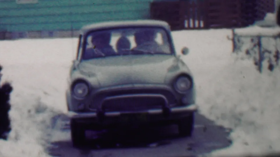 The 1958 home-movie frame shows a rounded light-colored car closer to camera, with bundled occupants visible through the windshield and snow piled around the driveway.