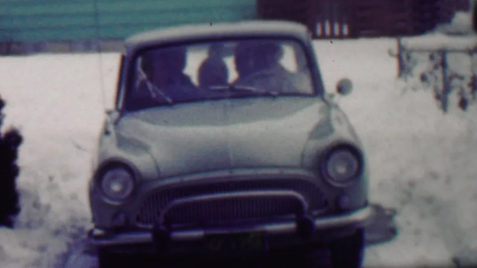 Close front view of a light-colored 1950s-style car on a snowy Grand Rapids driveway, with several passengers visible in soft focus through the windshield.