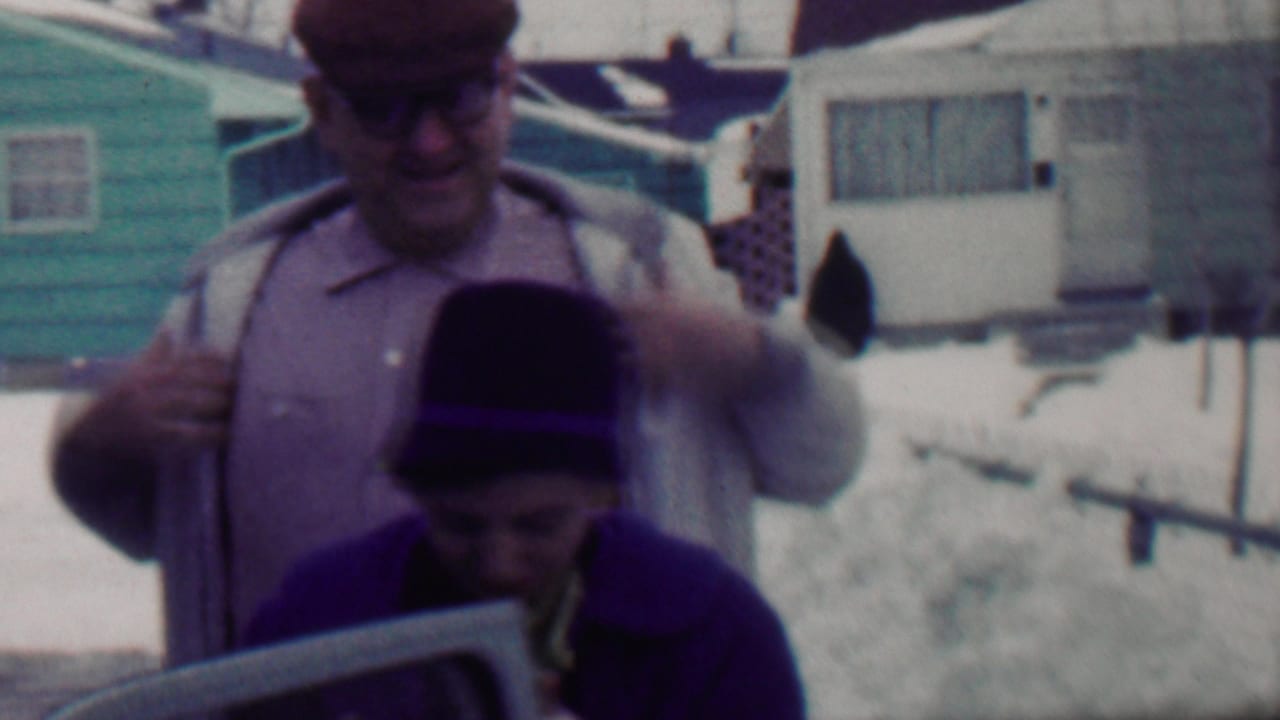In Grand Rapids, Michigan, 1958, a man in glasses and a light jacket stands behind a woman wearing a dark hat and purple coat beside a car with snowbanks and houses behind them.