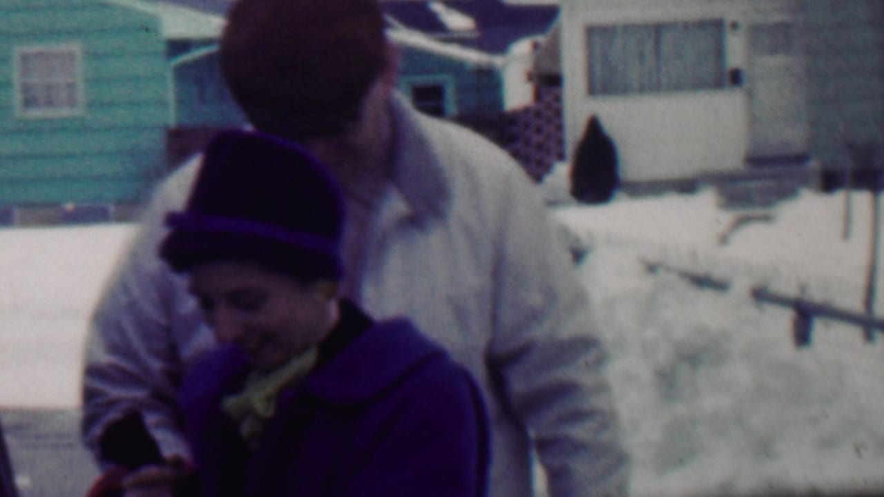 Man leans toward a woman in a dark hat as she looks down in a snowy residential setting.
