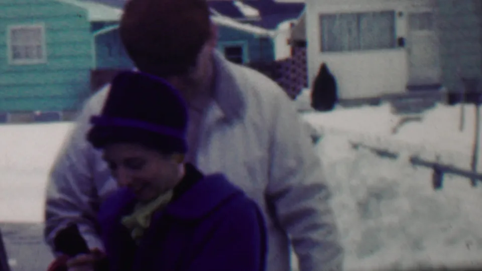 A man in a light jacket leans close behind a woman in a dark hat and purple coat, apparently helping adjust her clothing in the snowy Grand Rapids neighborhood setting.