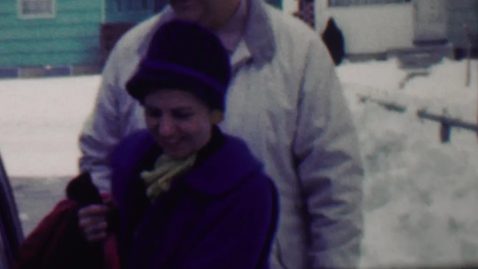 The woman stands in front of the man while holding something near her coat, with snowbanks, a car edge, and colorful residential houses visible behind them in the 1958 home-movie frame.