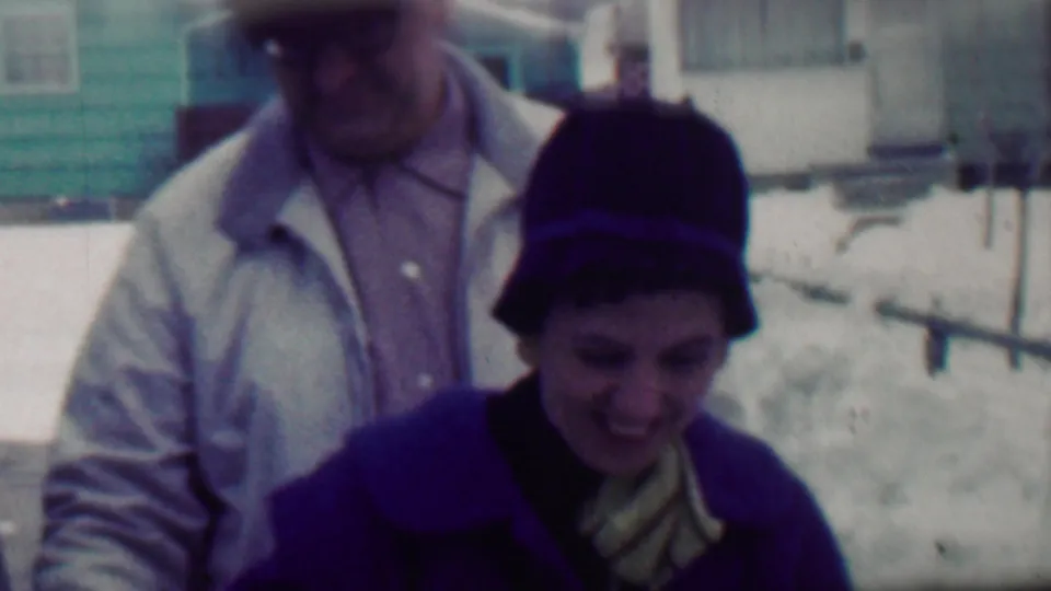 A smiling woman in a dark hat and purple coat turns slightly in front of a man wearing a light jacket, with snow-covered yards and residential houses in the background.