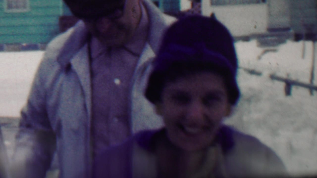 In this 1958 Grand Rapids home-movie frame, two bundled adults appear near a house in a snowy residential setting, with the foreground face soft and partly shadowed.