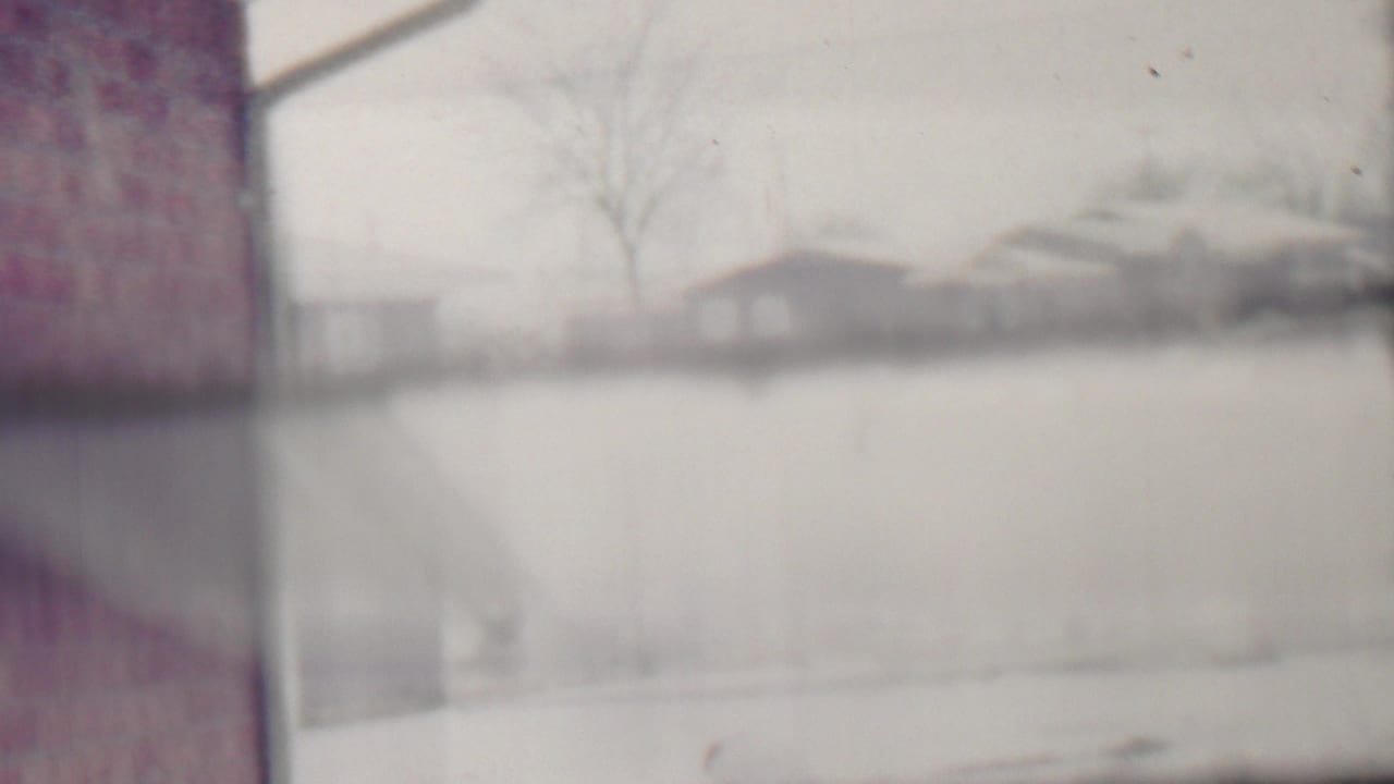 Snow-covered residential street with houses, bare trees, and a brick wall at the left edge.