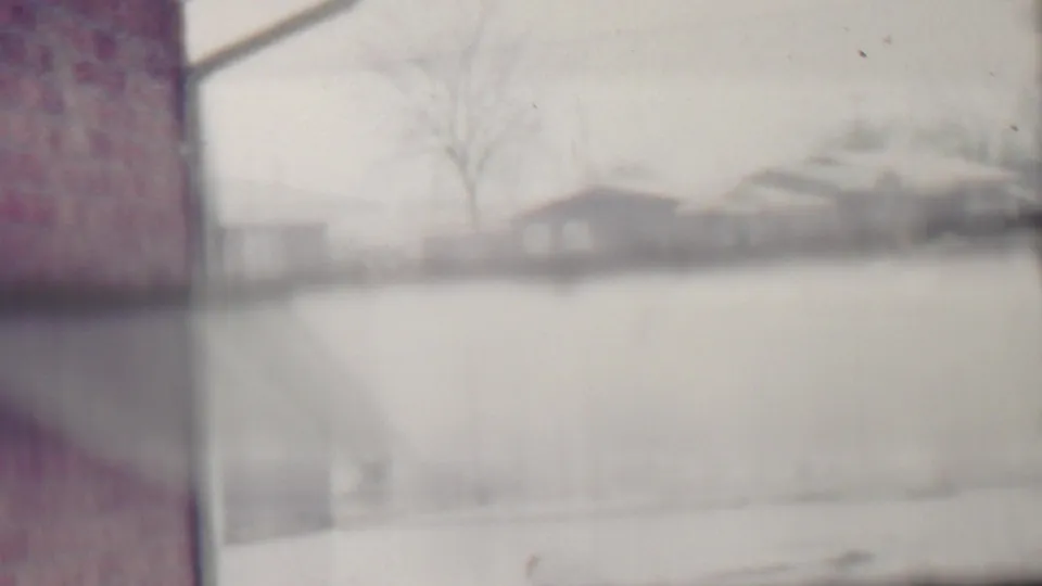 A snowy Grand Rapids residential street appears in soft focus, with modest houses, bare winter trees, and a brick wall or chimney along the left side of the frame.