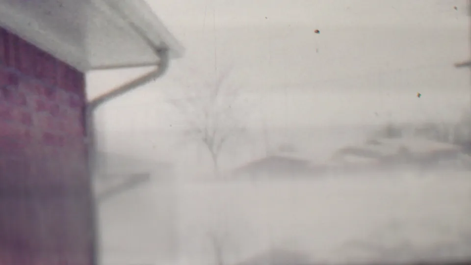 The frame shows a snow-covered residential view in Grand Rapids, with a house edge, brick wall or chimney, bare tree, and distant homes softened by 8mm focus and exposure.