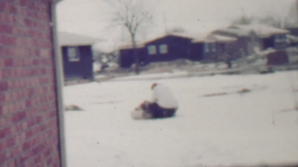 A snowy Grand Rapids neighborhood frame shows dark houses, a parked car, and a bundled figure low in the snow, possibly playing or sitting near the street.
