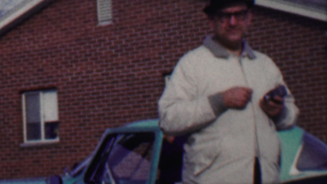 In supplied 1958 Grand Rapids home-movie footage, an older man in a dark fedora, glasses, and light jacket stands near a green car with a red brick building behind him.