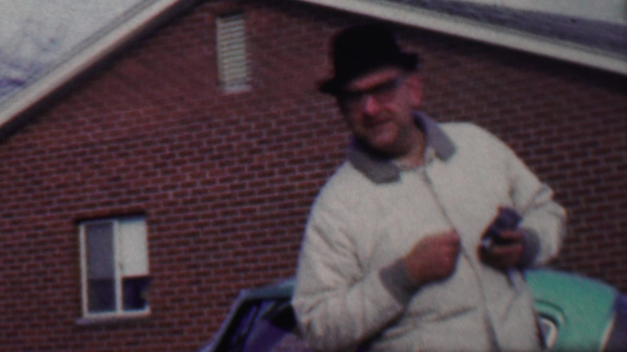 Man in fedora holds a small object while standing near a green car and brick wall.