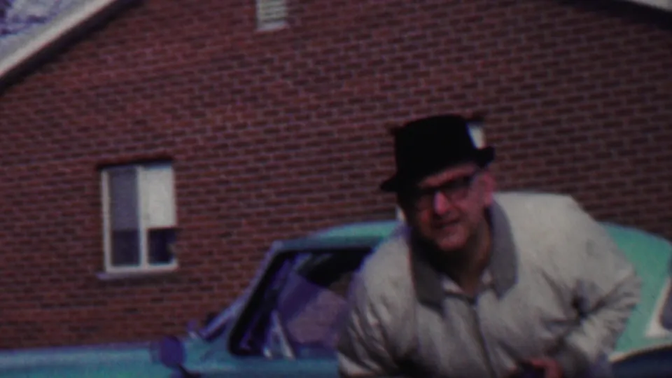 The man leans forward toward the camera, bringing his face closer while the green car and red brick building remain in the background.