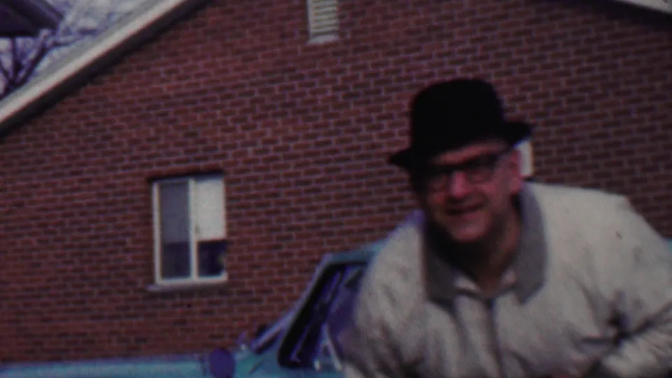 A closer handheld frame shows the man in fedora and glasses leaning into view, with the green car and brick building still partially visible behind him.