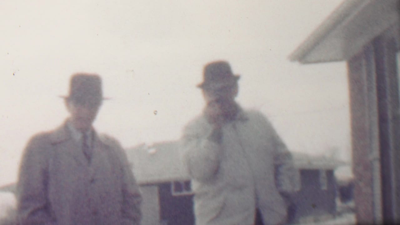 In metadata-supported 1958 Grand Rapids, two adult men in fedora-style hats and overcoats stand outdoors in a snowy residential setting, with one wearing a tie and the other holding a hand near his face.