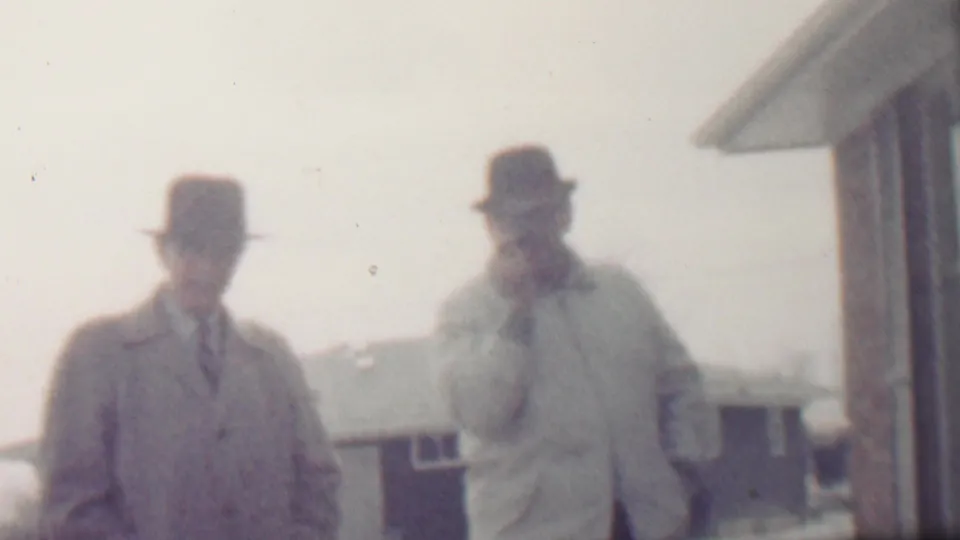 In metadata-supported 1958 Grand Rapids, two adult men in fedora-style hats and overcoats stand outdoors in a snowy residential setting, with one wearing a tie and the other holding a hand near his face.