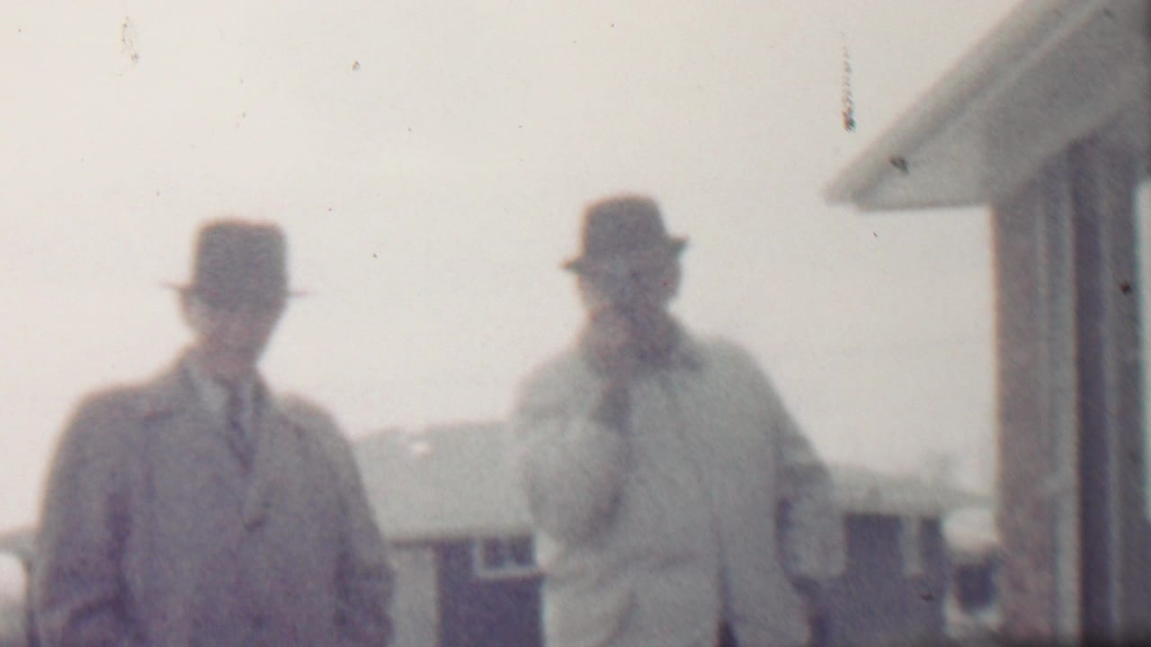 Two men in overcoats and hats stand beside a house in a snowy outdoor scene.