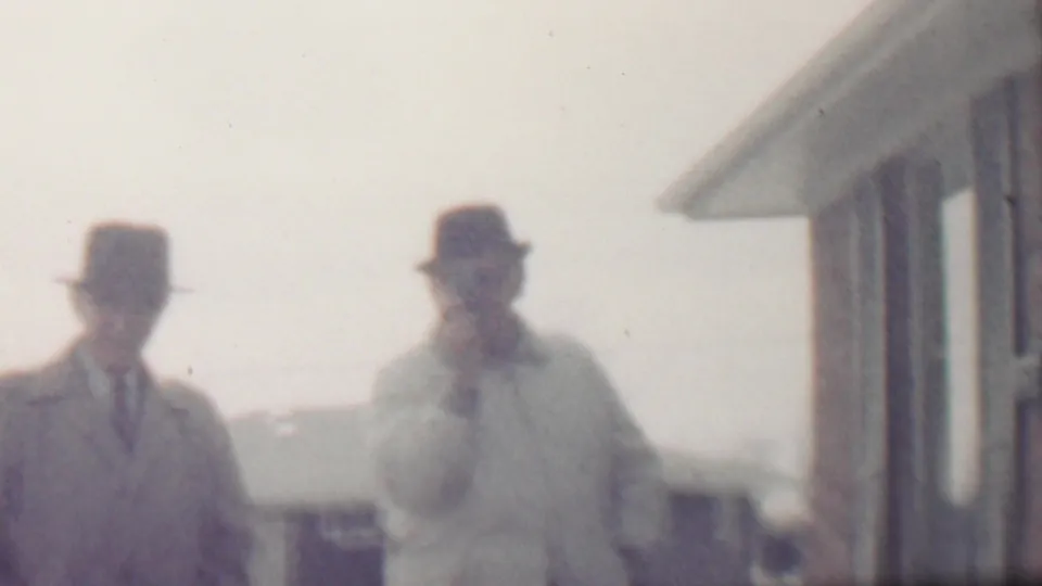 A soft 8mm frame shows two adult men in winter coats and hats standing in a residential snowy scene in the metadata-supported Grand Rapids location, with house exteriors visible behind them.