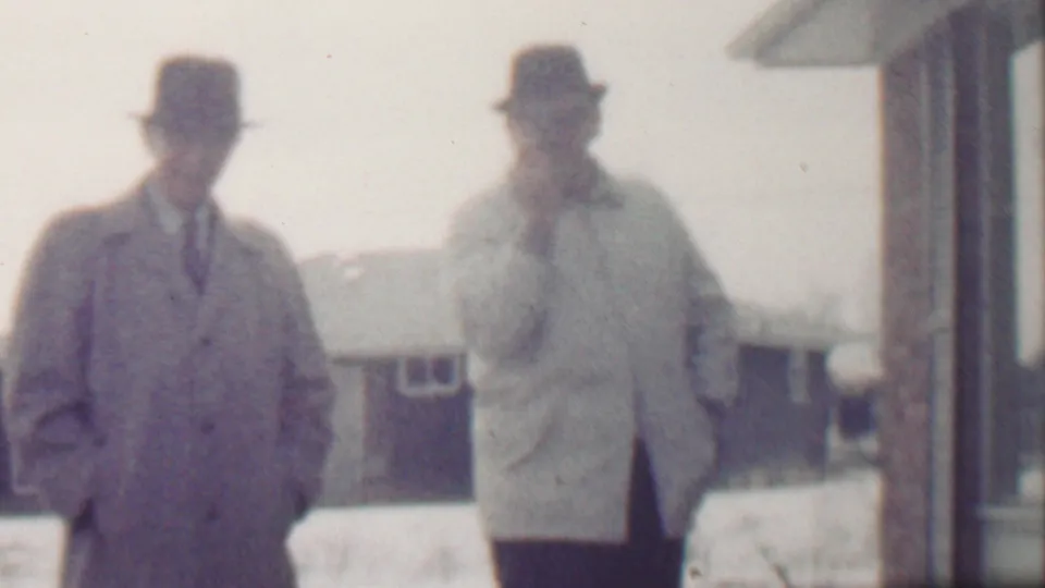 Two adult men in fedora-style hats and mid-century overcoats stand in a snowy residential exterior, with one man in a darker coat and tie and the other in a light coat near the house wall.