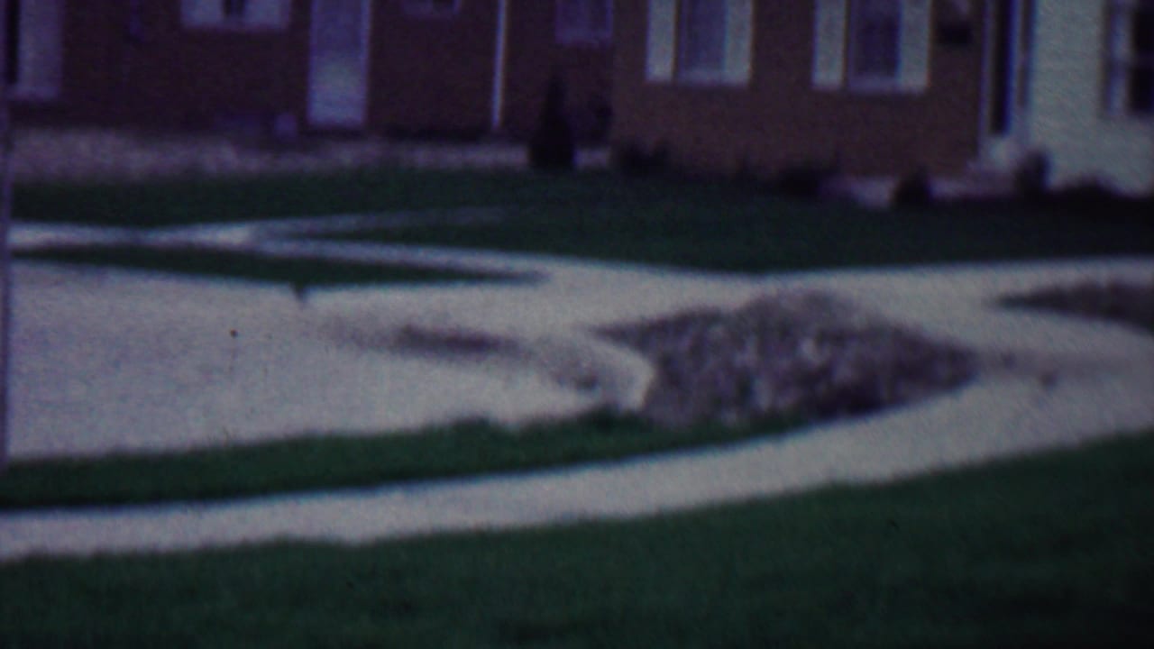 1958 Grand Rapids home-movie frame showing a pale concrete path across a green lawn with a brick house and white-trim windows in the background.