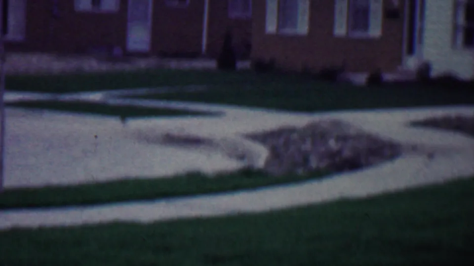1958 Grand Rapids home-movie frame showing a pale concrete path across a green lawn with a brick house and white-trim windows in the background.