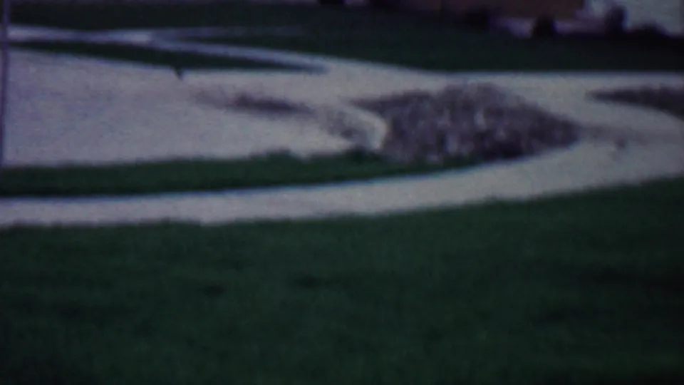 A soft 8mm frame from Grand Rapids, Michigan, showing a curved concrete walkway, green lawn, rubble pile, and partial brick residence in a 1958 suburban setting.