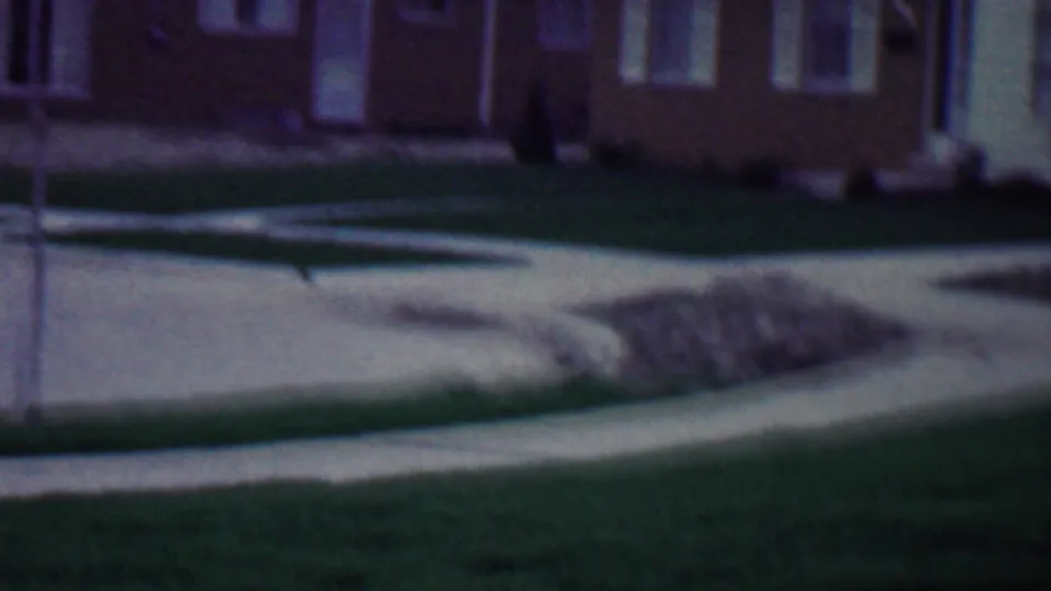 Grand Rapids 1958 home-movie frame with a brick house facade above a green lawn, a pale concrete path, and a small pile of construction debris near the path.