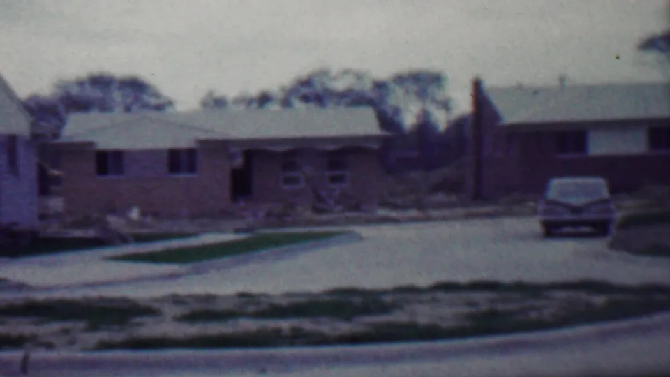 1958 home-movie frame of a Grand Rapids suburban block, showing a brick ranch house, driveway, curb, patchy grass, and a parked mid-century car.