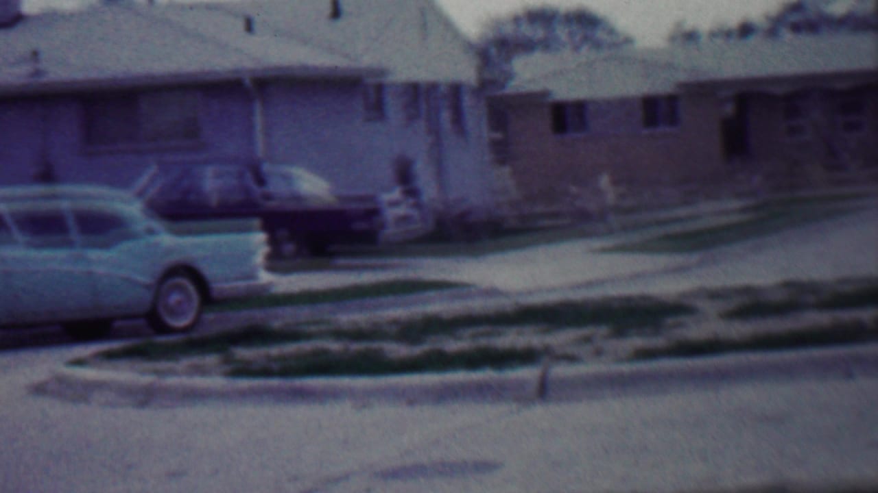 1958 Grand Rapids, Michigan home-movie frame showing parked finned cars beside single-story suburban houses with lawns and a paved street.